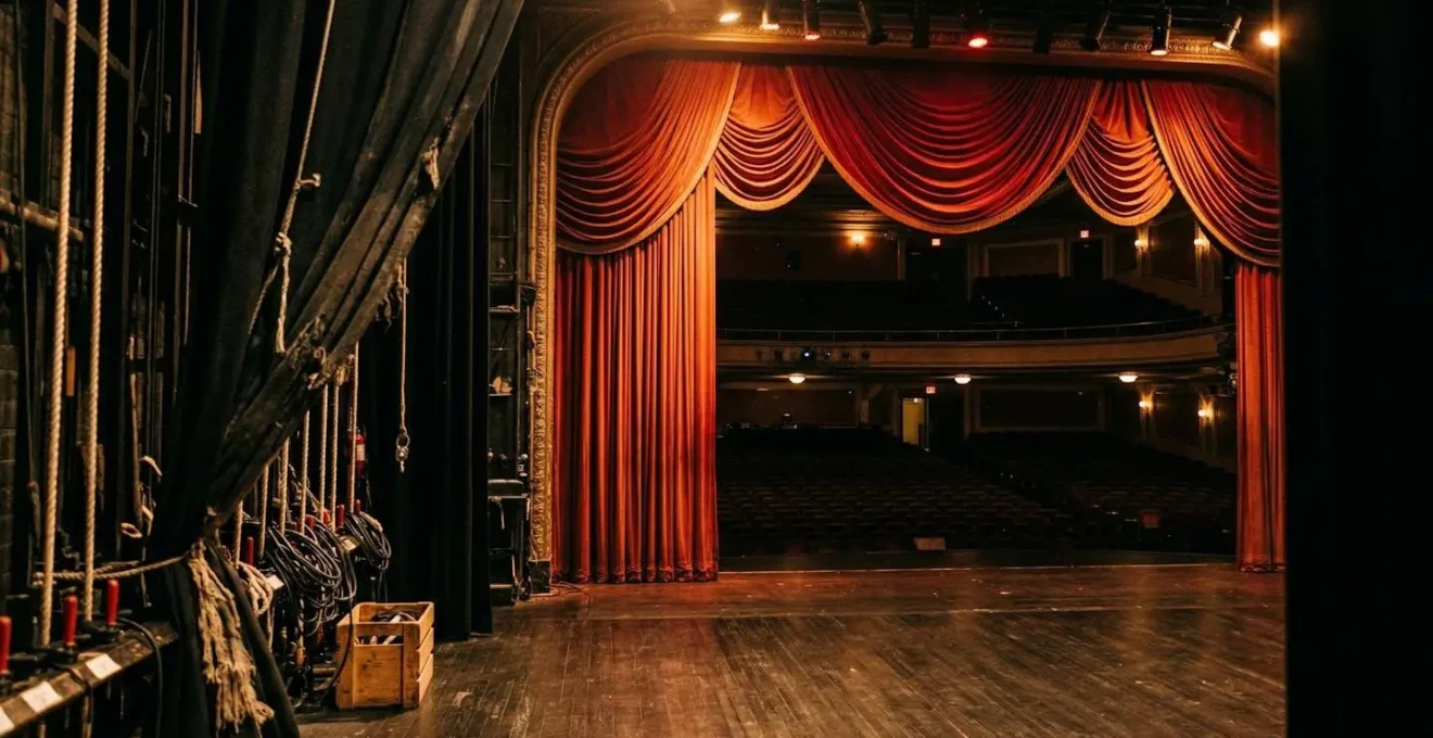 Vue depuis les coulisses d'une scène de théâtre vide baignée d'une lumière dramatique chaude avec des rideaux rouges en arrière-plan